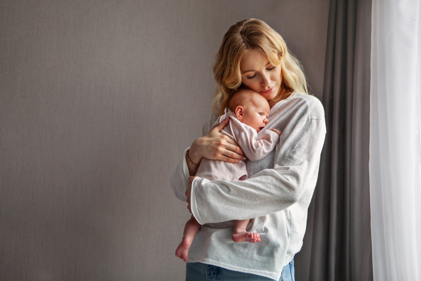 A mother standing by a window, cradling her sleeping newborn against her chest